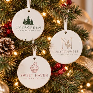May include: Three round white ceramic ornaments hanging from a Christmas tree. One features a green pine tree graphic and the text Evergreen Landscaping. Another shows a beige letter N with a leaf and Northwell Medical. One displays a pink cupcake and Sweet Haven Bakery.