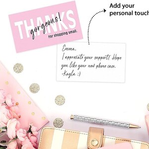 以下が含まれることがあります： ピンクのサンキューカードに「THANKS gorgeous! for shopping small.」というテキストと、「Emma. I appreciate your support! Hope you like your new phone case. -Kayla :)」という手書きのメモが書かれています