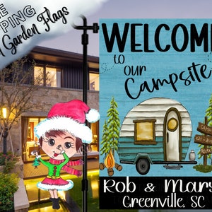 May include: A garden flag with the text "WELCOME to our Campsite" and a vintage camper illustration. The flag also includes the names "Rob & Mary" and "Greenville, SC". The flag is displayed outdoors.
