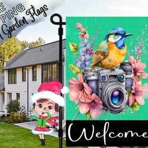 May include: A teal green garden flag with a watercolor illustration of a blue and yellow bird perched on a vintage camera. Pink and purple flowers surround the camera. The text "Welcome" is printed in black at the bottom of the flag.