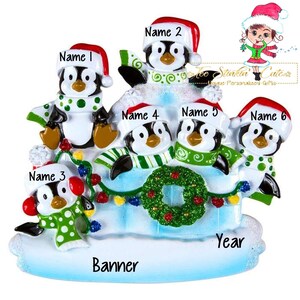 Puede incluir: Un adorno navideño con seis pingüinos en un iglú cubierto de nieve con una corona verde y luces. Los pingüinos llevan gorros de Papá Noel y bufandas. El adorno tiene una pancarta que dice "Banner" y un espacio para el año.