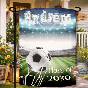 May include: A personalized garden flag featuring a soccer ball in a net against a stadium backdrop. The flag displays the name "Andrew" in bold letters, with "class of 2030" below. The design incorporates green, white, and blue hues.