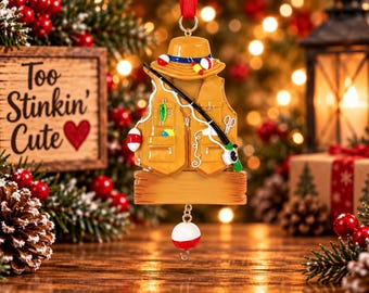 Könnte beinhalten: Ein hängender Weihnachtsbaumschmuck in Form einer beigen Angelweste mit passendem Hut und Angelrute. Die Weste ist mit bunten Ködern und einem Schwimmer verziert. Unten hängt ein leeres Holzschild, das an einem roten Band befestigt ist.
