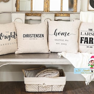 May include: Four decorative throw pillows with personalized text on a light beige fabric. The pillows are arranged on a metal bench with a white blanket draped over the end. The pillows feature the text "The Wrights Farmhouse", "Christensen", "home Seattle, WA Est. 2003", and "The Larsen Farm Est. 2001".