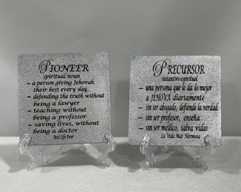 May include: Two silver glitter plaques with black text on stands. The left plaque reads "Pioneer" and defines the word as a spiritual noun. The right plaque reads "Precursor" and defines the word as a spiritual noun in Spanish.