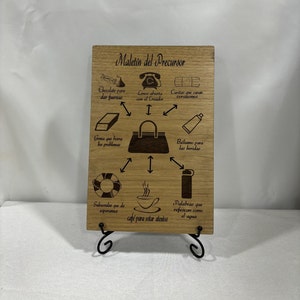 Peut inclure: Une plaque en bois avec le texte "Maletin del Precursor" et un schéma d'une mallette avec divers objets autour. Les objets comprennent du chocolat, un téléphone, des bandages, une gomme, un tube de pommade, une bouée de sauvetage, une tasse de café et une bouteille d'eau. Le texte autour des objets décrit leur but, comme "Chocolate para dar fuerzas" (Chocolat pour donner de la force) et "Goma que borra los problemas" (Gomme qui efface les problèmes).