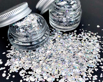 Könnte beinhalten: Drei kleine Gläser gefüllt mit silbernem Glitzer, mit leicht geöffneten Deckeln. Der Glitzer ist um die Gläser herum auf einer schwarzen Oberfläche verstreut. Auf den Gläsern steht der Text "ecoGlimmer".