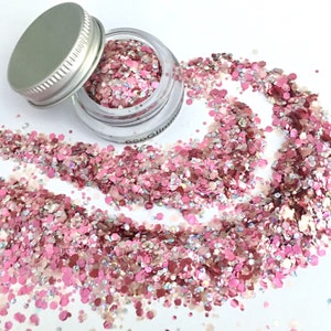 May include: A small glass jar filled with pink, white, and iridescent glitter. The jar has a silver lid and is open, with glitter spilling out onto a white surface.