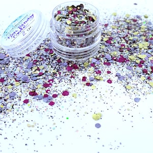 May include: A small clear jar filled with a mix of purple, gold, and silver glitter. The glitter is scattered around the jar on a white surface. The jar has a label that reads "Eco Glitter Non-Fussy Fuchsia 2 gr."