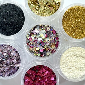 May include: Six small jars filled with different colors of glitter. The jars are arranged in a circle, with the glitter colors visible in each jar. The glitter colors are black, gold, pink, white, purple, and a mix of pink, gold, and white.