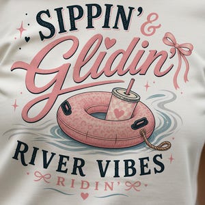 May include: A cream-colored t-shirt featuring a graphic print with the text "Sippin' & Glidin' RIVER VIBES RIDIN'". The design includes a pink leopard-print inner tube floating on water, a cup with a straw, a pink bow, and small star accents.