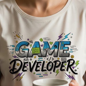 May include: Cream-colored t-shirt with the words "GAME DEVELOPER" in a colorful, illustrated design. The design includes game controllers, lightning bolts, and stars. A person is holding a white mug.