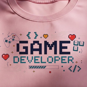 May include: A light pink sweatshirt with the words "GAME DEVELOPER" in pixelated navy blue letters. The design includes a game controller, hearts, and coding symbols. The sweatshirt is folded neatly.