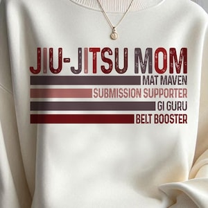 May include: White sweatshirt with the text "Jiu-Jitsu Mom" in red and black letters. The text is layered with colorful stripes and includes the words "Mat Maven", "Submission Supporter", "Gi Guru", and "Belt Booster".