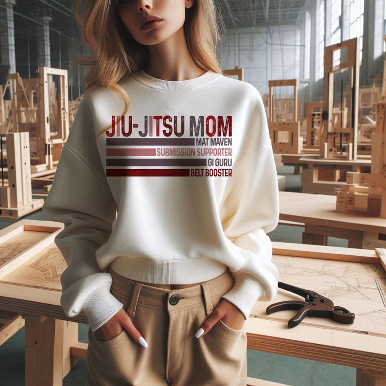 May include: White sweatshirt with the text "Jiu-Jitsu Mom" and the words "Mat Maven", "Submission Supporter", "Gi Guru", and "Belt Booster" printed in red and brown on a striped background.