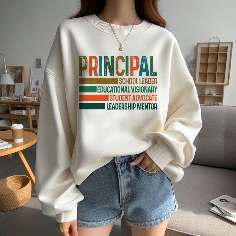 K&ouml;nnte beinhalten: Wei&szlig;er Pullover mit dem Text "PRINCIPAL" in Gr&uuml;n, Orange und Braun. Der Text ist von einem gr&uuml;n-orange-braunen Streifenrand umgeben. Der Text unter "PRINCIPAL" lautet "SCHOOL LEADER", "EDUCATIONAL VISIONARY", "STUDENT ADVOCATE" und "LEADERSHIP MENTOR".