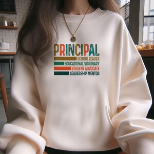 K&ouml;nnte beinhalten: Ein wei&szlig;er Pullover mit dem Wort "PRINCIPAL" in bunten Retro-Lettern. Der Text darunter lautet "Schulleiter, Bildungsvision&auml;r, Sch&uuml;leranwalt, F&uuml;hrungsmentor."