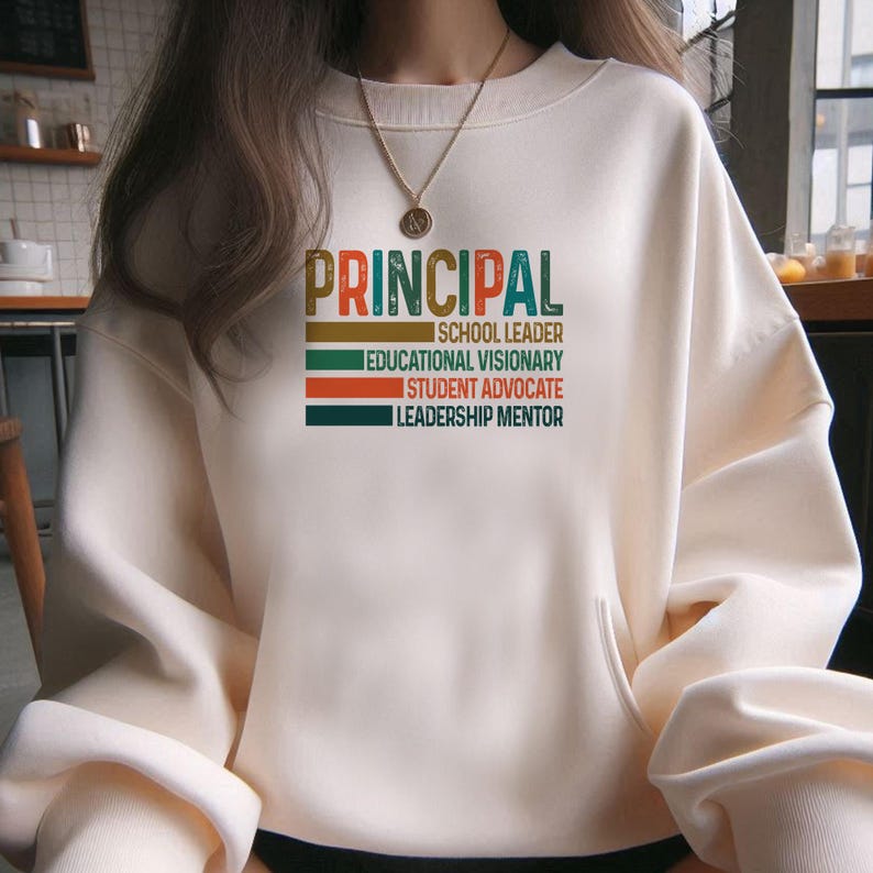 K&ouml;nnte beinhalten: Ein wei&szlig;er Pullover mit dem Wort "PRINCIPAL" in bunten Retro-Lettern. Der Text darunter lautet "Schulleiter, Bildungsvision&auml;r, Sch&uuml;leranwalt, F&uuml;hrungsmentor."