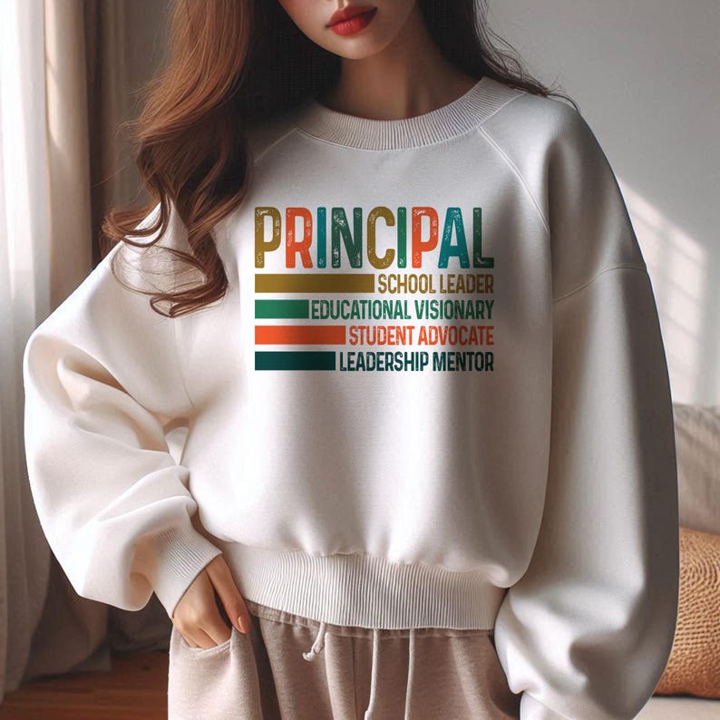 K&ouml;nnte beinhalten: Wei&szlig;er Pullover mit dem Text "PRINCIPAL" in einer Retro-Schriftart mit bunten Streifen darunter. Der Text lautet "Schulleiter, Bildungsvision&auml;r, Sch&uuml;leranwalt, F&uuml;hrungsmentor."