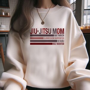 May include: White sweatshirt with the text "Jiu-Jitsu Mom" in black and red. The text is layered with the words "Mat Maven", "Submission Supporter", "Gi Guru", and "Belt Booster" in smaller font.