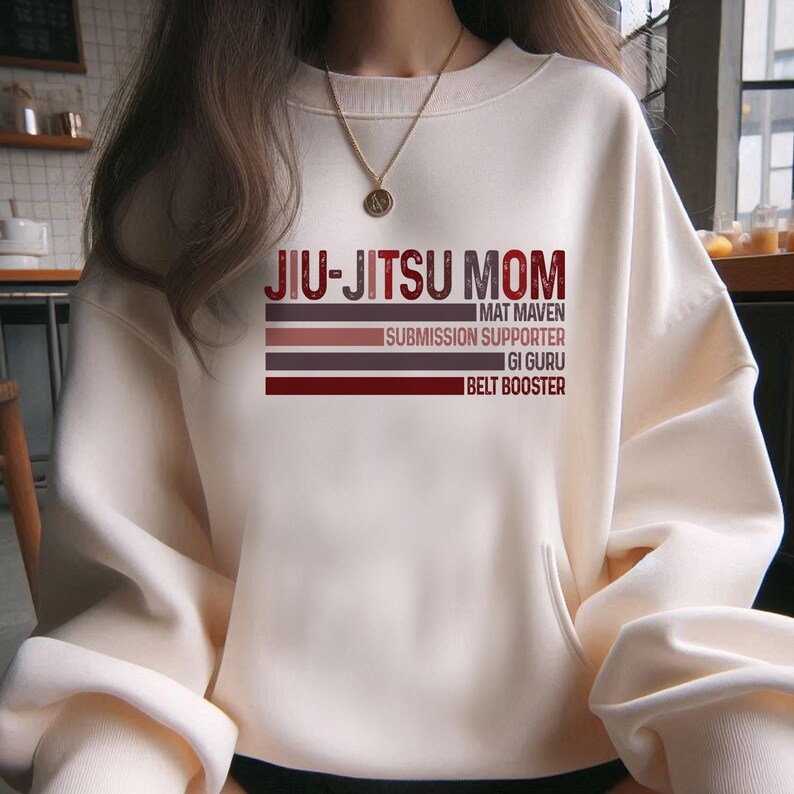 May include: White sweatshirt with the text "Jiu-Jitsu Mom" in black and red. The text is layered with the words "Mat Maven", "Submission Supporter", "Gi Guru", and "Belt Booster" in smaller font.