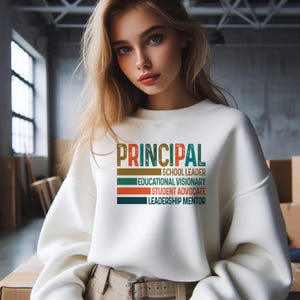 K&ouml;nnte beinhalten: Wei&szlig;er Pullover mit dem Text "PRINCIPAL" in bunten Buchstaben. Der Text wird von einer Liste von Berufsbezeichnungen gefolgt: "Schulleiter", "Bildungsvision&auml;r", "Sch&uuml;leranwalt" und "F&uuml;hrungsmentor".