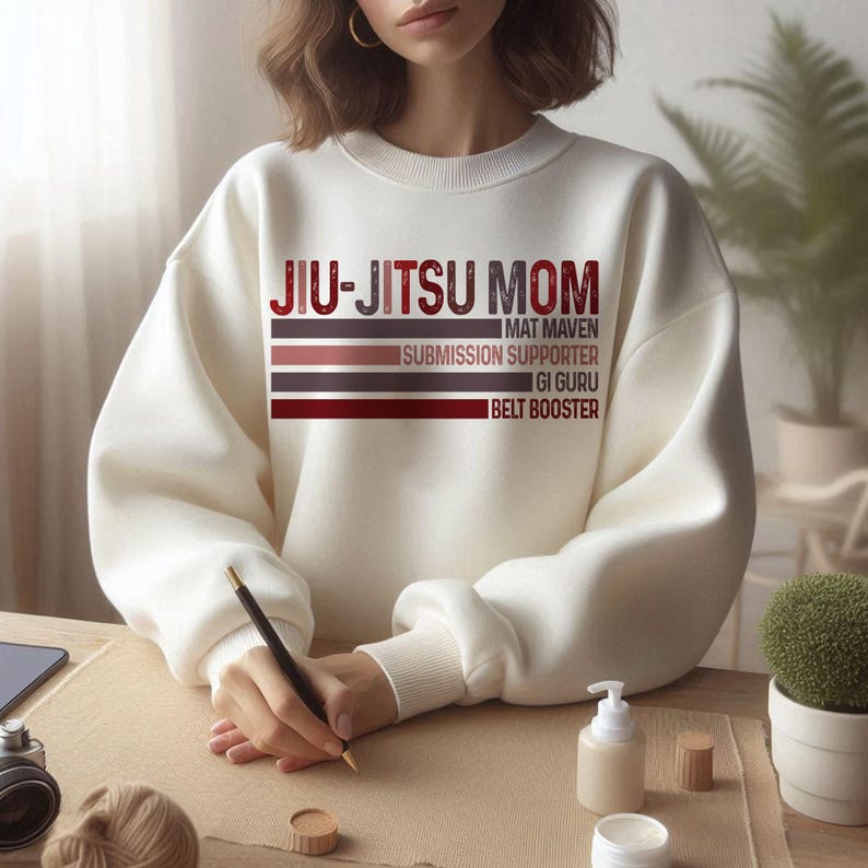 May include: White sweatshirt with the text "Jiu-Jitsu Mom" in red and brown lettering. The text is layered with the words "Mat Maven", "Submission Supporter", "Gi Guru", and "Belt Booster" underneath.