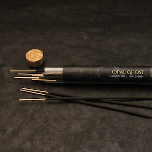 May include: A black tube of incense sticks with the text "OPAL GHOST" and the scent description "Moonflower Lemon Juniper." Several incense sticks are visible, along with a cork stopper. The background is a dark, textured surface.