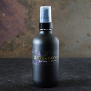May include: A black glass bottle with a black spray top and a white label that says "Sleeper Choir Black Tea, Fig, Bergamot".