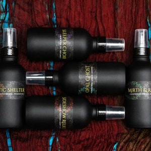 May include: Five black glass bottles with spray tops and labels. The labels have names like "Sleeper Choir", "Opal Ghost", "Sideshow Relic", "Poetic Shelter", and "Mirth & Ritual". Each label also includes a description of the scent.