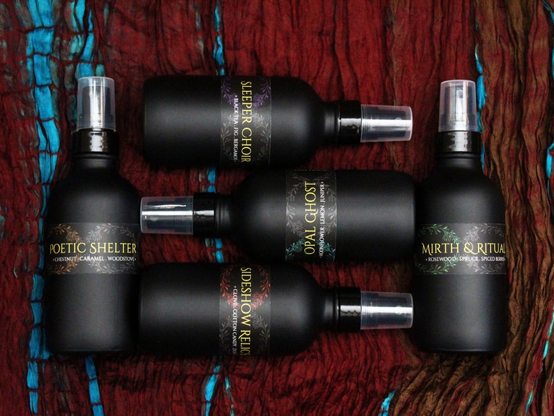 May include: Five black glass bottles with spray tops and labels. The labels have names like "Sleeper Choir", "Opal Ghost", "Sideshow Relic", "Poetic Shelter", and "Mirth & Ritual". Each label also includes a description of the scent.