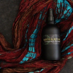 May include: A black glass bottle with a spray nozzle, labeled "Mirth & Ritual - Rosewood, Spruce, Spiced Berries". The bottle is sitting on a red and blue patterned fabric.