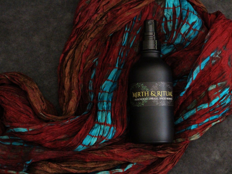 May include: A black glass bottle with a spray nozzle, labeled "Mirth & Ritual - Rosewood, Spruce, Spiced Berries". The bottle is sitting on a red and blue patterned fabric.