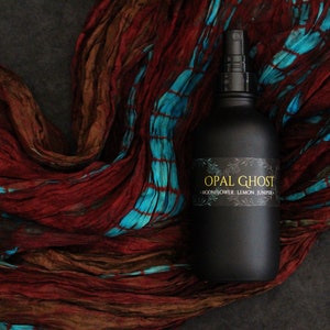 May include: A black glass bottle with a spray nozzle, labeled "OPAL GHOST - MOONFLOWER, LEMON, JUNIPER". The bottle is sitting on a red and blue patterned fabric.