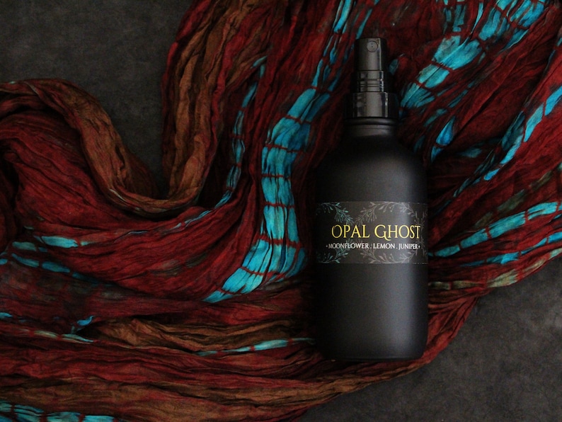 May include: A black glass bottle with a spray nozzle, labeled "OPAL GHOST - MOONFLOWER, LEMON, JUNIPER". The bottle is sitting on a red and blue patterned fabric.
