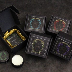 May include: Five black square boxes containing solid perfume, each with a unique label featuring a botanical wreath and text including Opal Ghost, Sideshow Relics, Starlight Kind, Sleeper Choir, and Grotto & Muse. Two small black round tins are included.