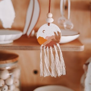 May include: A ceramic ornament with a painted landscape scene of mountains, a crescent moon, and stars. The ornament is attached to a brown leather cord with a wooden bead and a white tassel.