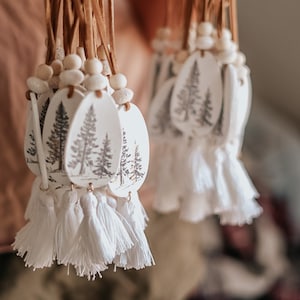 May include: A collection of white ceramic ornaments with a brown leather cord and white tassels. Each ornament features a black and white illustration of a pine tree.