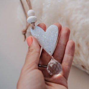 May include: A white ceramic heart with a sparkly finish hangs from a brown leather cord with white beads. A clear crystal prism dangles from the bottom of the heart.