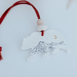 May include: White ceramic ornament shaped like a bear with a mountain scene and a crescent moon. The ornament has a red cord and a wooden bead.