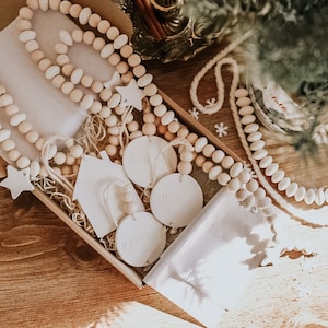 May include: A set of wooden bead garlands with white ceramic ornaments in the shape of stars and houses. The ornaments are blank and can be personalized. The garlands are packaged in a brown box with white tissue paper.