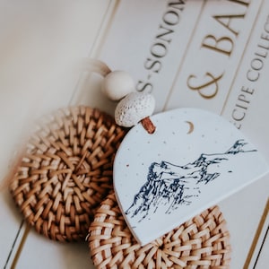 May include: A white ceramic ornament with a black and white mountain scene and a crescent moon. The ornament is hanging from a string of white beads and is resting on two woven rattan coasters.