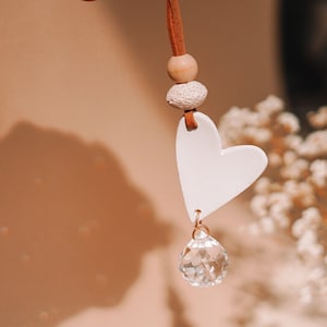 May include: A white ceramic heart charm with a crystal pendant hanging from a brown leather cord with wooden beads.