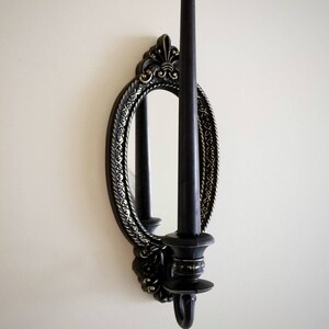 May include: Black candle holder with an oval mirror and ornate gold detailing. The candle holder is mounted on a white wall.