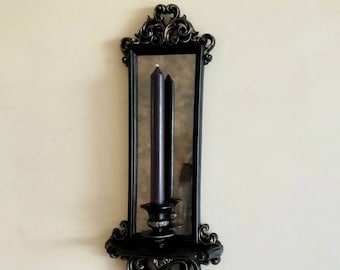 May include: Black ornate wall-mounted candle holder with a mirror and a single black candle. The holder has a decorative design with scrolls and a shelf.