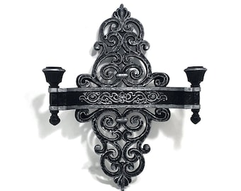 May include: A black metal wall sconce with a decorative scroll design. The sconce has two candle holders and a shelf.