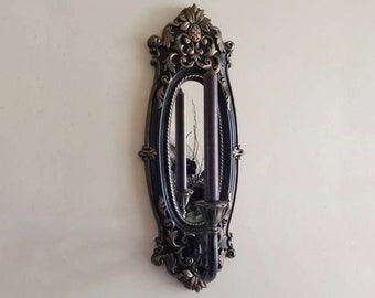 May include: An ornate, dark-colored wall sconce with a mirrored oval center and two tall, dark candles. The sconce has a decorative, antique-style design with gold accents and is mounted on a light-colored wall.