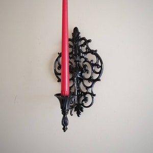 Puede incluir: Candelabro de pared de metal negro con un diseño ornamentado. Una vela roja se coloca en el candelabro.