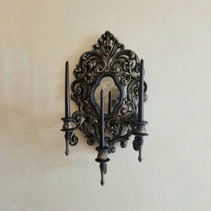 Puede incluir: Una pared de aplique de pared negro y dorado con un espejo y cuatro candelabros. El aplique tiene un diseño decorativo con detalles intrincados.