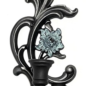 May include: A black metal wall sconce with a single candle holder. The sconce features a decorative floral design with a blue and white flower.
